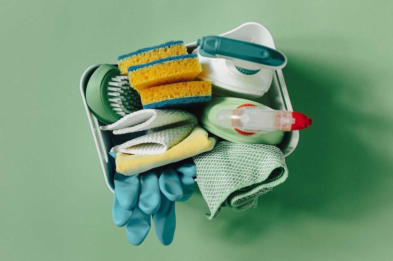 Een witte mand gevuld met schoonmaakartikelen zoals sponzen, microvezeldoeken, een borstel, spuitflessen en rubberen handschoenen.