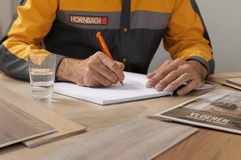 Hornbach medewerker met Hornbach Logo plant een vloerproject aan een tafel met vloerstalen, een brochure en een notitieblok.
