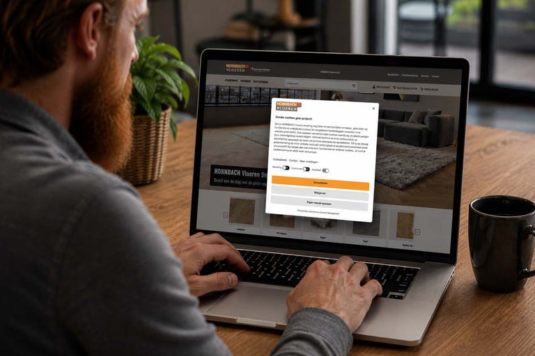 Een man aan een houten tafel gebruikt een laptop met de Hornbach Vloeren website en een cookiebanner.
