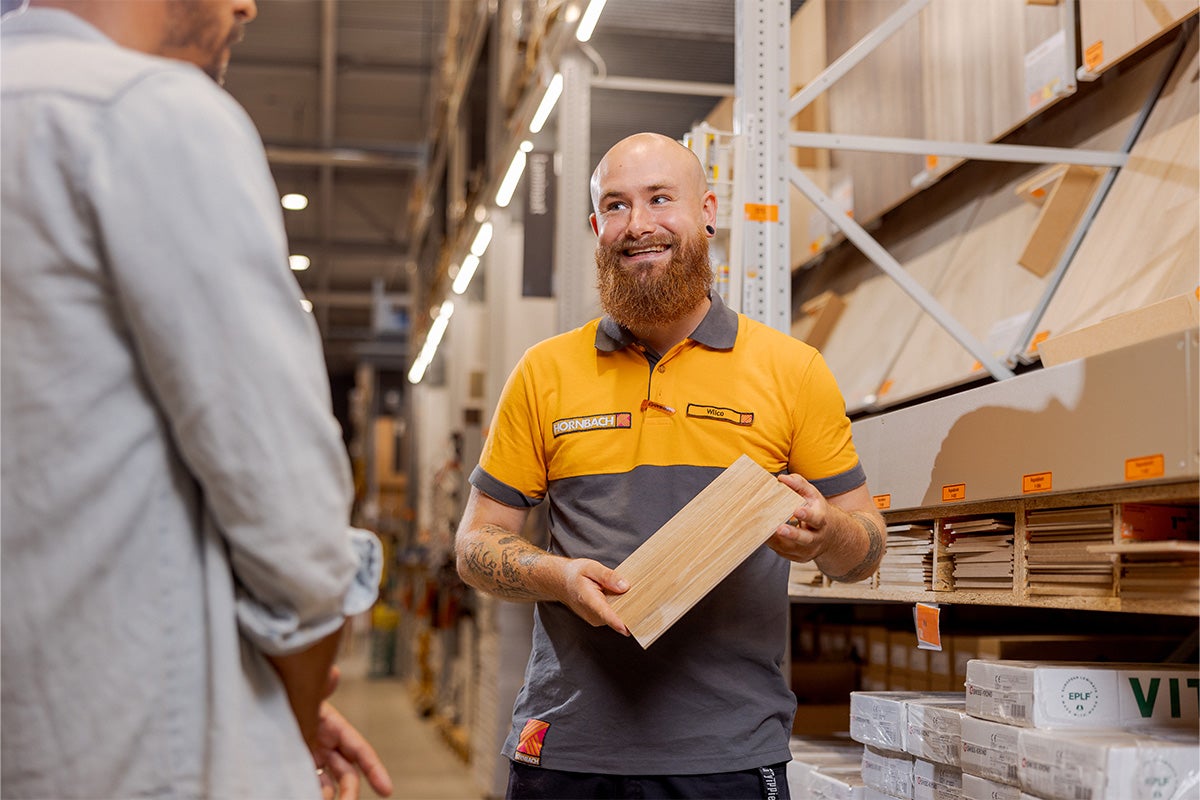 Een Hornbach medewerker in werkkleding adviseert een klant in de bouwmarkt en houdt een houten vloerstaal vast.