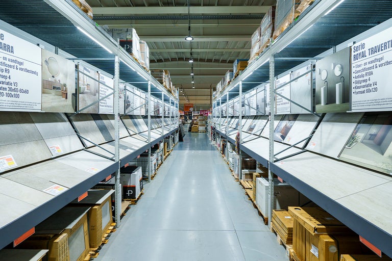 Helder verlicht gangpad in een bouwmarkt met grootformaat wand- en vloertegels in hoge metalen rekken.