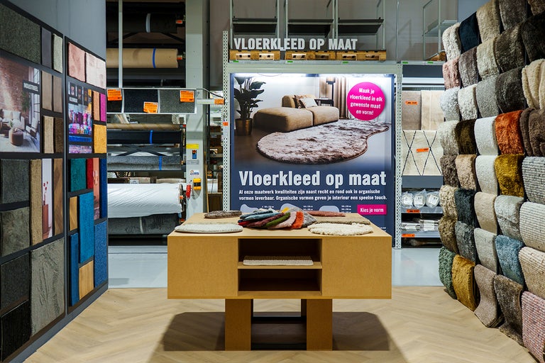 Lichte tapijtafdeling met centrale presentatietafel voor stalen, poster voor vloerkleden op maat en diverse wand- en rekdisplays met tapijten.