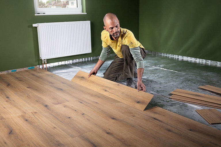 Een man legt laminaat met eikenlook op een zilveren ondervloer in een kamer met groene muren en een radiator.