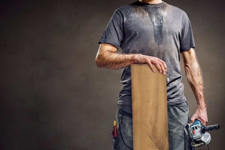 Een man in werkkleding houdt een houten vloerplank en een haakse slijper vast.