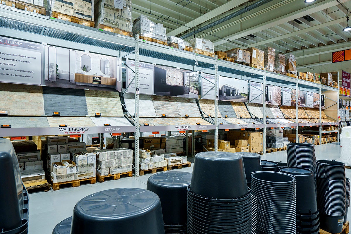 Een georganiseerd gangpad in de Hornbach bouwmarkt met wandbekleding in het schap, pallets met goederen en zwarte emmers op de voorgrond.