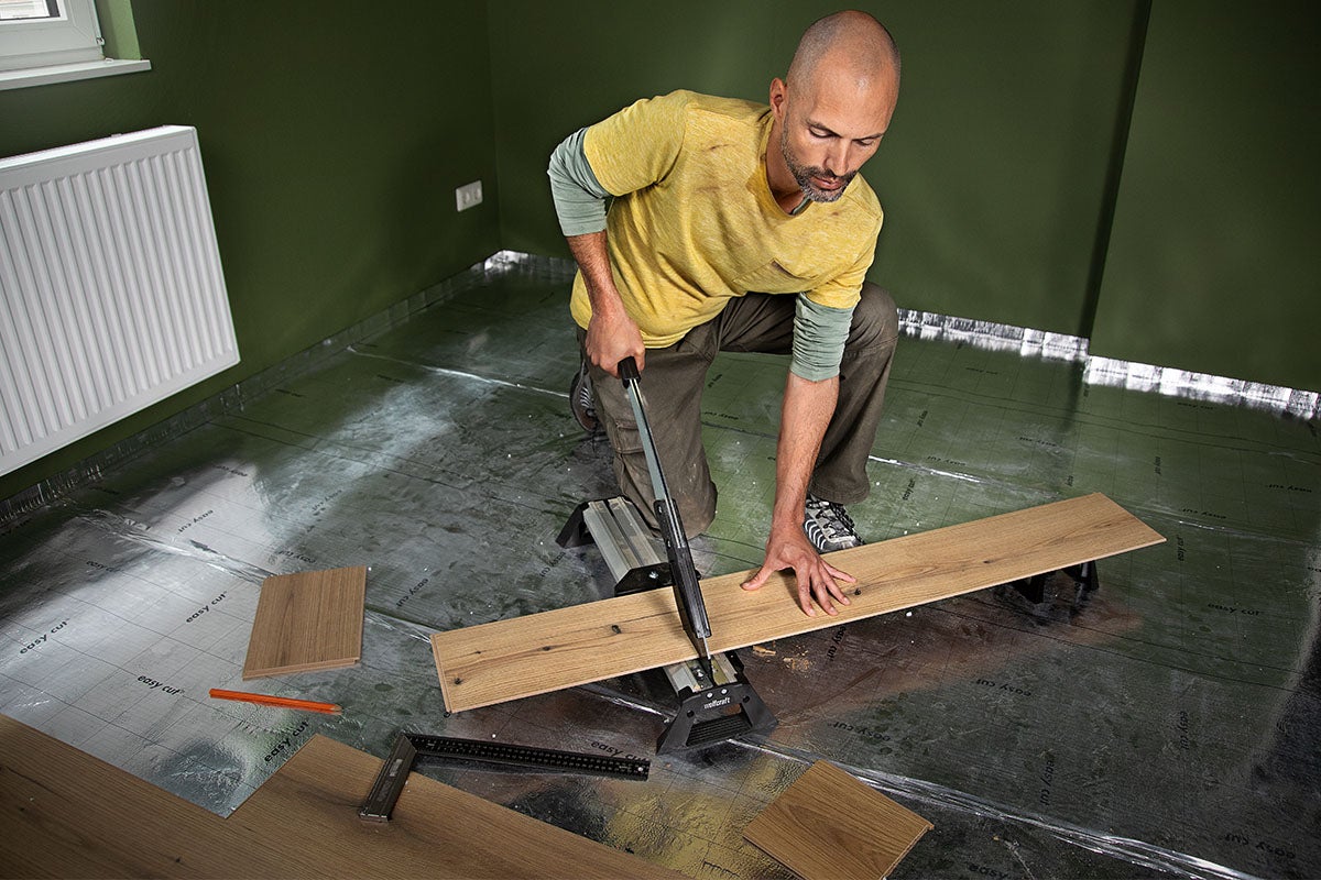 Een man snijdt een laminaatplank met een laminaatsnijder op een zilveren ondervloer in een kamer met groene muren.