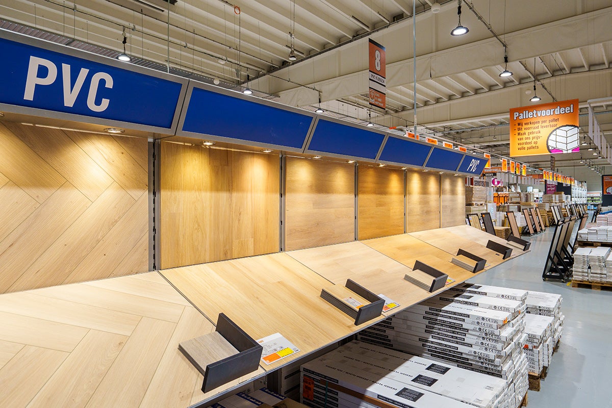 Grote presentatie van PVC-vloeren in een bouwmarkt met diverse lichte houtpatronen zoals visgraat en brede planken op een stelling.