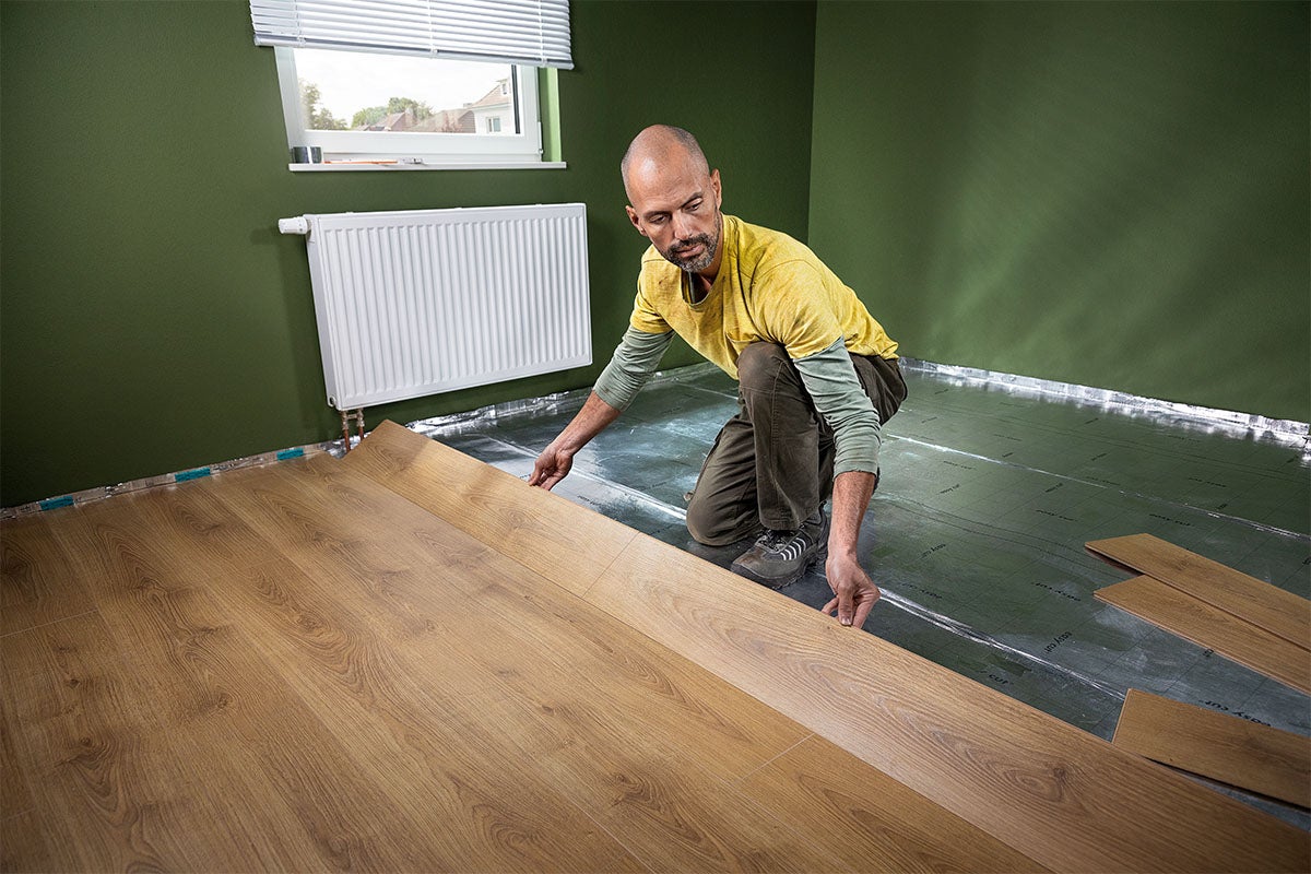 Een man legt een laminaatvloer met houtlook op een ondervloer in een ruimte met een radiator.