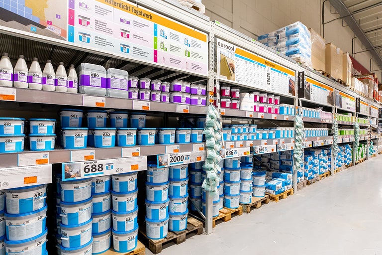 Lichte, georganiseerde magazijngang bij Hornbach met metalen rekken vol Eurocol tegelproducten en Eurocol Logo op informatieborden.