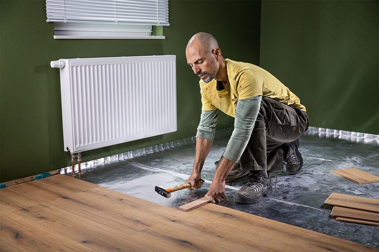 Man legt laminaatvloer met hamer en aanslagblok in een lichte kamer met radiator en ondervloer.