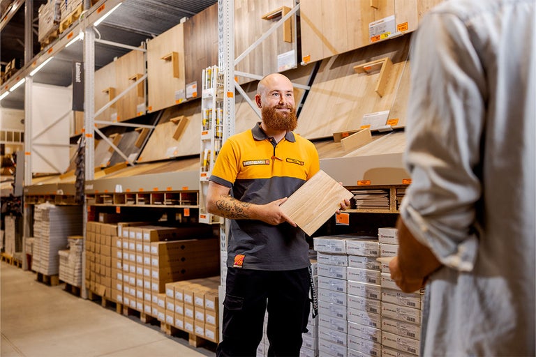 Hornbach medewerker adviseert klant op de georganiseerde vloerenafdeling en houdt een laminaatstaal vast voor stellingen met diverse vloeren.
