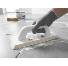 Een persoon met een werkhandschoen brengt met een epoxy voegspaan wit voegmiddel aan op grijze vloertegels.