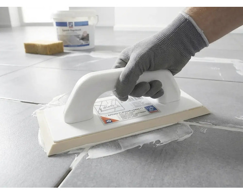Een persoon met een werkhandschoen brengt met een epoxy voegspaan wit voegmiddel aan op grijze vloertegels.