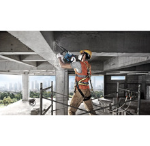 Bouwvakker met beschermende uitrusting gebruikt Bosch boorhamer op een betonstructuur op een bouwplaats met steiger en stadsgezicht.