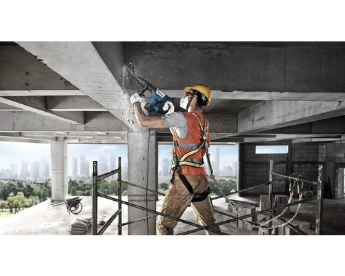 Bouwvakker met beschermende uitrusting gebruikt Bosch boorhamer op een betonstructuur op een bouwplaats met steiger en stadsgezicht.