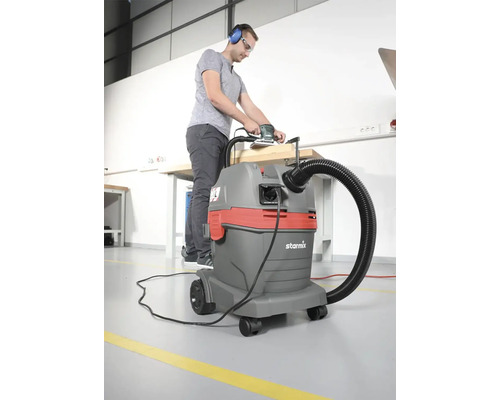 In een lichte werkplaats gebruikt een man een schuurmachine die is aangesloten op een Starmix industriële stofzuiger aan een werkbank.