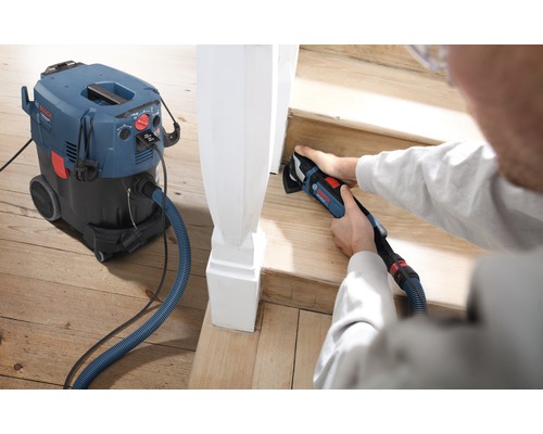 Een persoon gebruikt een multifunctionele schuurmachine om een houten trap te schuren, met een stofzuiger voor een schone werkomgeving.