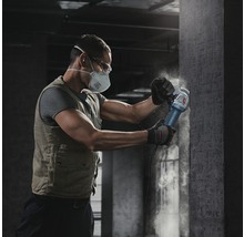 Een man met een beschermend masker en veiligheidsbril werkt met een haakse slijper op een betonnen muur.