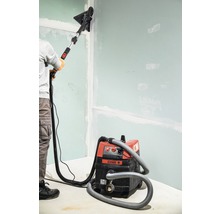 Een persoon schuurt een muur met een gipsplaatschuurmachine en een stofzuiger.
