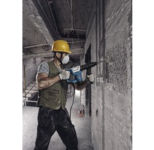 Een bouwvakker met veiligheidshelm en stofmasker gebruikt een boorhamer op een betonnen muur.