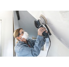 Een vrouw schuurt een plafond met een excentrische schuurmachine en draagt een veiligheidsmasker en een veiligheidsbril.