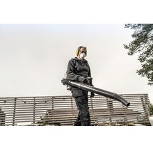 Een persoon bedient een ruggedragen bladblazer in de tuin, beschermd met gehoorbescherming en een stofmasker.