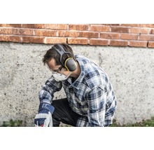 Een man knielt en draagt persoonlijke beschermingsmiddelen, waaronder gehoorbescherming, een veiligheidsbril, een stofmasker en werkhandschoenen.