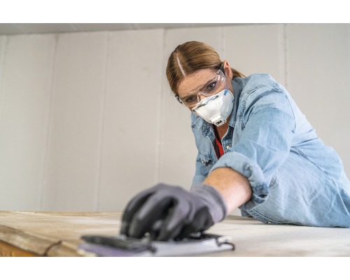 Vrouw schuurt hout met een schuurblok en draagt een veiligheidsbril en stofmasker