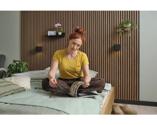 Een vrouw zit op een bed voor een wandbekleding en leest een tijdschrift. De slaapkamer is licht en vriendelijk ingericht.
