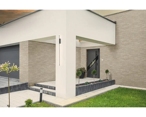 Moderne zwarte buitenwandlamp op een witte pilaar bij de entree van een woning met bakstenen gevel en garage.