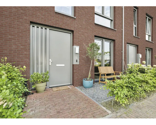 Moderne entree met grijze voordeur, bakstenen gevel, houten bankje op grind en groene beplanting.