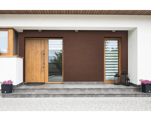 Moderne huisingang met donkerbruine bakstenen gevel, houten deur, grote ramen, grijze tegeltrappen en bloembakken.
