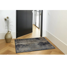 Rechthoekige deurmat met abstract patroon op houten parket in een lichte hal naast een zwarte deur en een decoratieve vaas.