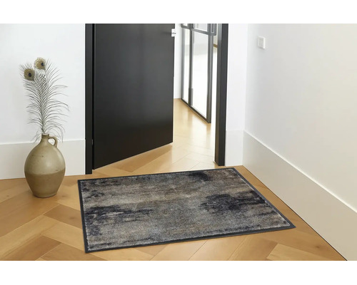 Rechthoekige deurmat met abstract patroon op houten parket in een lichte hal naast een zwarte deur en een decoratieve vaas.