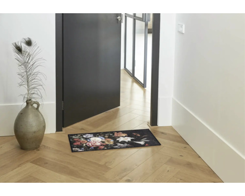 Deurmat met bloemenpatroon in een lichte hal met houten vloer naast een deur en een vaas.