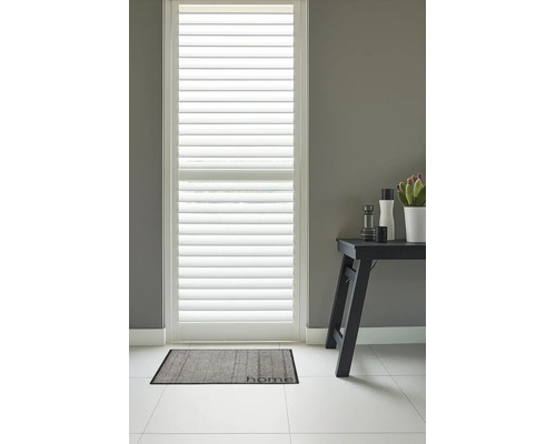 Witte houten shutters voor een raam tegen een grijze muur met een grijze deurmat met de tekst home op een lichte tegelvloer.