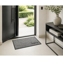 Lichte, moderne entree met een open voordeur en een gemusterde deurmat met het woord home. Rechts staat een zwarte sidetable.