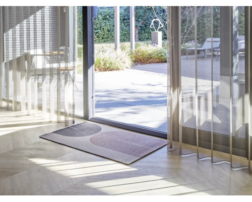 Moderne deurmat met geometrisch boogpatroon op een lichte houten vloer voor een glazen terrasdeur met transparante gordijnen.