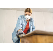 Vrouw schuurt een meubelstuk met schuurpapier, ze draagt een beschermend masker en een veiligheidsbril