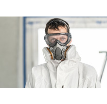 Een persoon draagt een veiligheidsbril, een ademhalingsmasker en beschermende kleding.