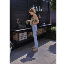 Een vrouw geeft planten water met een gieter op een terras met planken, houten kisten en een vloermat.
