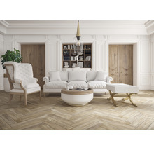 Woonkamer met bank, fauteuil, voetenbank, salontafel en boekenkast in een lichte kamer.