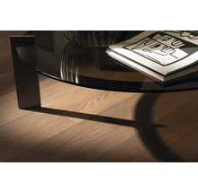 Detail van een ronde salontafel met glazen blad en tijdschriften op een houten vloer.