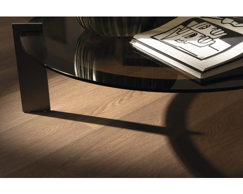 Detail van een ronde salontafel met glazen blad en tijdschriften op een houten vloer.